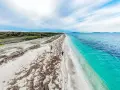 Una persona cammina su una spiaggia di sabbia bianca, con alghe sparse, verso un mare turchese e profondo. Un paesaggio ezzi mannu.