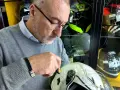 Uomo con occhiali regola un casco da moto bianco, mentre altri caschi sono esposti. Si nota l'anno produzione casco moto su un'etichetta.