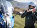 Uomo in giacca di pelle che indossa caschi per moto bianchi, pronto per un viaggio su una moto blu sfocata.
