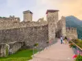 Un sentiero fiorito conduce al maestoso Castello San Michele, con le sue torri imponenti e mura antiche che si stagliano contro il cielo.