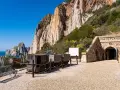 Antichi vagoni minerari e l'ingresso di Porto Flavia, testimoni della storica zona mineraria sarda, si stagliano contro un imponente promontorio roccioso.