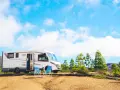Un camper bianco è parcheggiato in un'area sterrata con due sedie blu e un tavolino. Sullo sfondo, pini e colline sotto un cielo azzurro con nuvole bianche. Un luogo ideale per un soggiorno in stile piscin'e janas.
