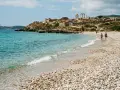 Spiaggia di ciottoli con rovine antiche in collina e un faro in lontananza. Una coppia passeggia sulla riva, godendosi la brezza marina. La facella cabras illumina il paesaggio.