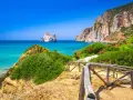 Sentiero con staccionata in legno conduce a una spiaggia con acqua cristallina e l'isolotto Pan di Zucchero in Sardegna.