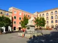La piazza Tola a Sassari, con il monumento al centro, edifici colorati e alberi verdi sotto un cielo azzurro.