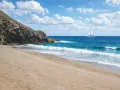 Spiaggia dorata e mare azzurro vicino a Sassari, con un catamarano bianco che naviga all'orizzonte sotto un cielo nuvoloso.
