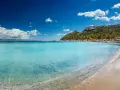 Acque cristalline e sabbia dorata sulle spiagge di Cagliari, con il promontorio verdeggiante e il porto turistico sullo sfondo.