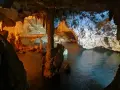 Interno di una grotta con stalattiti e stalagmiti illuminate da luci arancioni e blu. L'acqua scura riflette le formazioni rocciose in questa grotta verde.