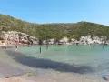 Persone si godono il sole e il mare cristallino nella spiaggia cala lunga, con colline verdi e rocce a fare da cornice.