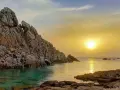 Scogliere rocciose bagnate da acque cristalline, con il sole che tramonta sulla valle della luna, creando riflessi dorati sul mare.