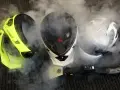 Tre caschi, uno giallo fluo, uno bianco e nero, e uno bianco, sono avvolti dal vapore. Un accessorio per la pulizia a vapore è posizionato davanti a loro, suggerendo che il casco non deve essere pulito con solventi.