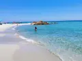 Spiaggia di Berchida: sabbia bianca, acque cristalline e rocce rosse sotto un cielo azzurro. Una donna si gode il mare calmo.