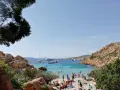 Spiaggia affollata a Cala Coticcio, con gente che prende il sole e nuota nelle acque cristalline. Diverse barche e yacht ancorati nella baia.