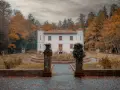 Un'antica villa bianca con torrette, circondata da alberi autunnali dai colori caldi. Un sentiero lastricato conduce all'ingresso, tra statue e vegetazione. Le **badde salighes** sussurrano storie di tempi passati.