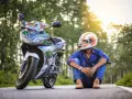 Giovane con casco da moto arancione e bianco, seduto accanto alla sua Yamaha. Spera di allargare il casco stretto per un viaggio più comodo.