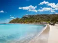 Spiaggia con acque cristalline e la sagoma della Sella del Diavolo all'orizzonte, sotto un cielo azzurro con nuvole bianche.