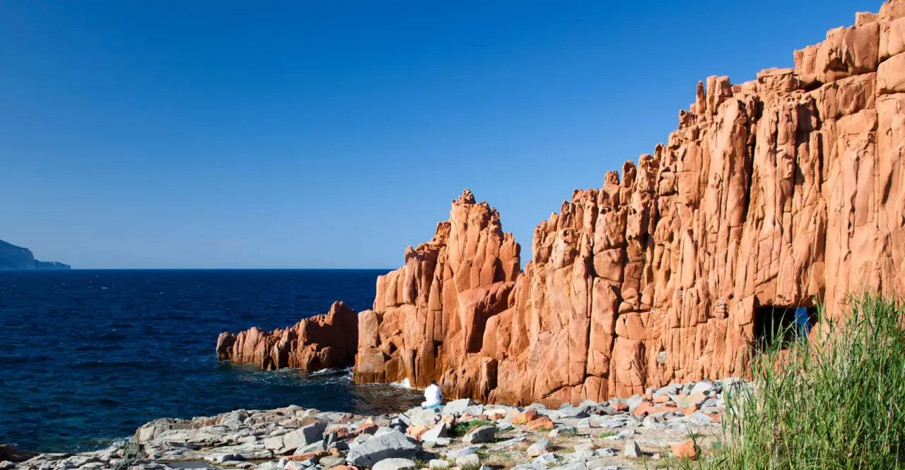 Le rocce rosse di Arbatax si ergono maestose sul mare blu. Una figura solitaria medita ai loro piedi, immersa nella bellezza selvaggia.