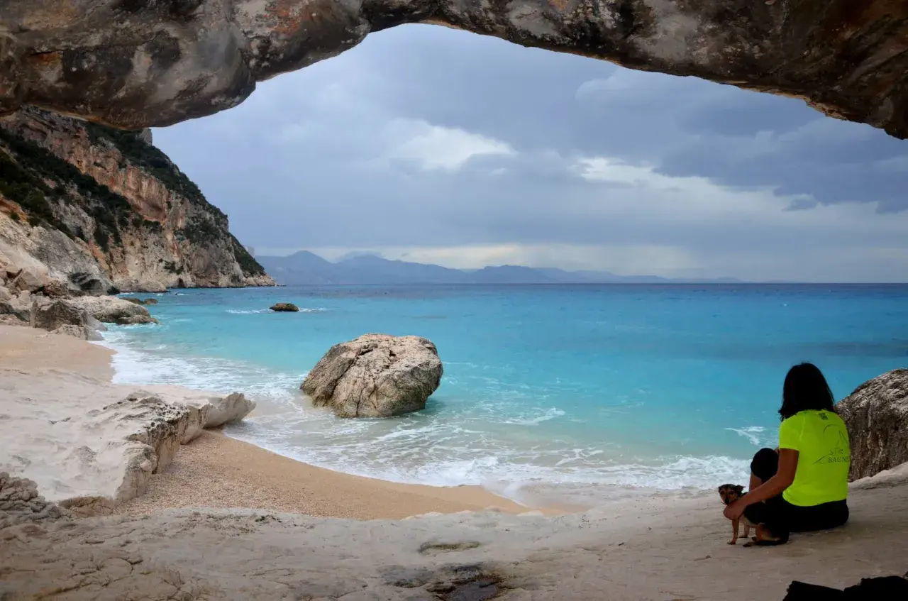 Vista da una grotta su Cala Goloritzé, con mare turchese, scogliere e una donna con un cane.