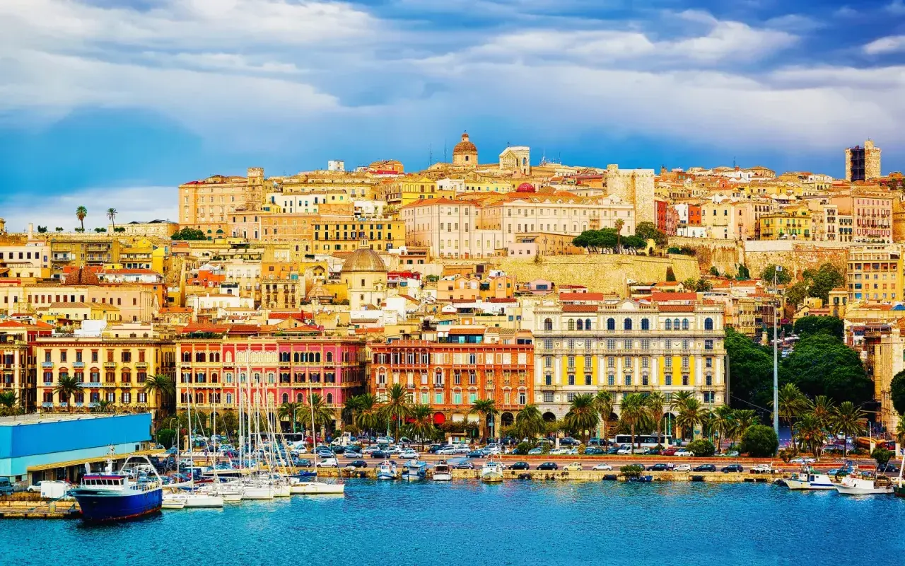 Panoramica colorata di **Mirrionis**, con edifici storici, un porto con barche a vela e un cielo azzurro con nuvole.