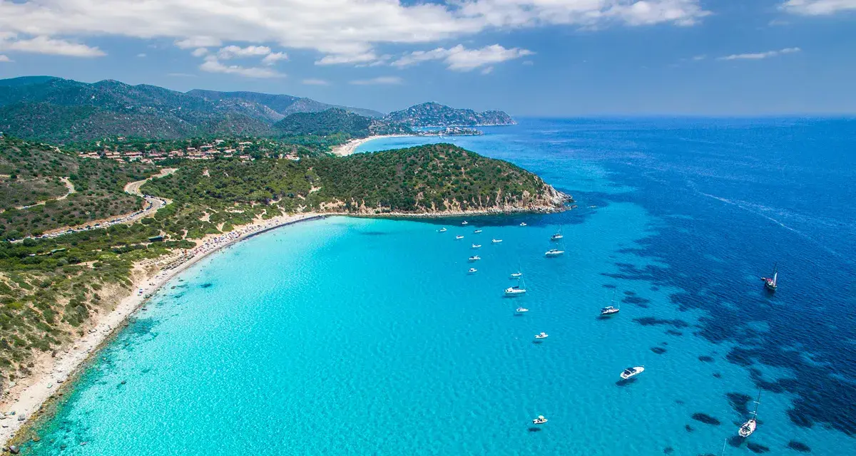 Spiaggia di Chia, Sardegna: acque cristalline che sembrano mari pintau, con barche a vela e un litorale affollato.