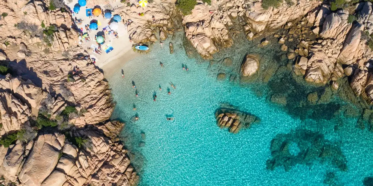 Vista aerea della spiaggia di Cala Coticcio, con bagnanti che si godono le acque cristalline e ombrelloni colorati sulla sabbia dorata.