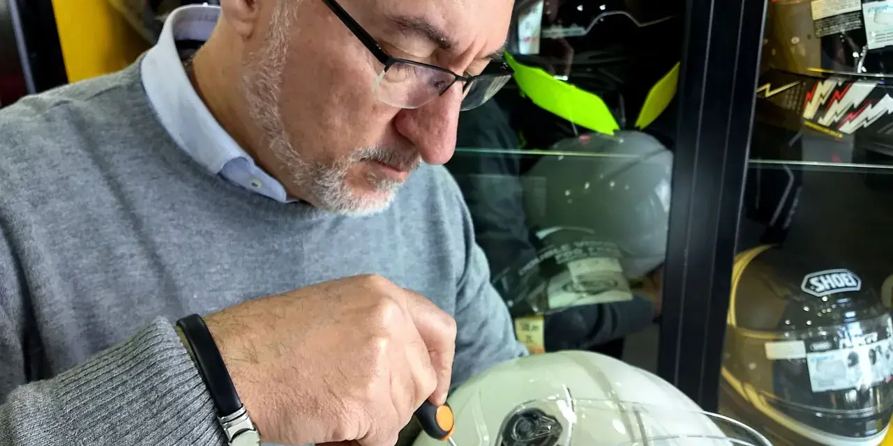 Uomo con occhiali regola un casco da moto bianco, mentre altri caschi sono esposti. Si nota l'anno produzione casco moto su un'etichetta.