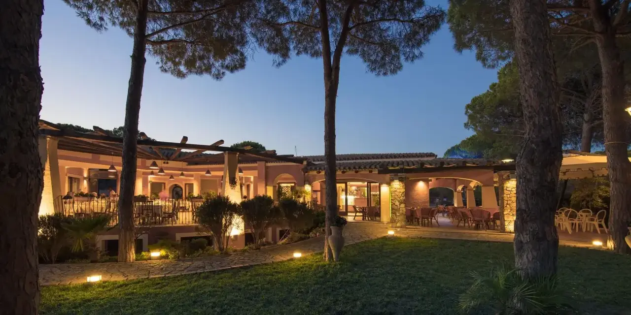 La sorgente Villasimius: un ristorante con tavoli all'aperto illuminati da luci soffuse, circondato da pini e un prato curato.