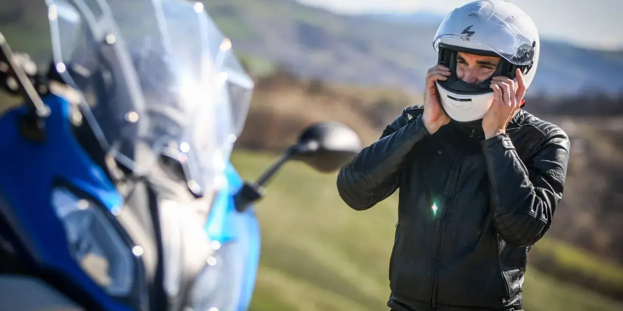 Uomo in giacca di pelle che indossa caschi per moto bianchi, pronto per un viaggio su una moto blu sfocata.
