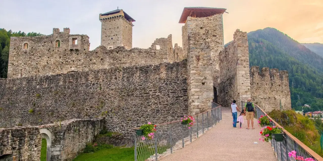 Un sentiero fiorito conduce al maestoso Castello San Michele, con le sue torri imponenti e mura antiche che si stagliano contro il cielo.