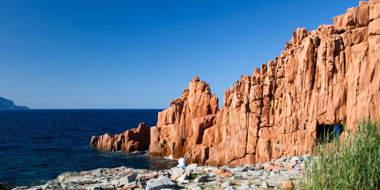 Le rocce rosse di Arbatax si ergono maestose sul mare blu. Una figura solitaria medita ai loro piedi, immersa nella bellezza selvaggia.
