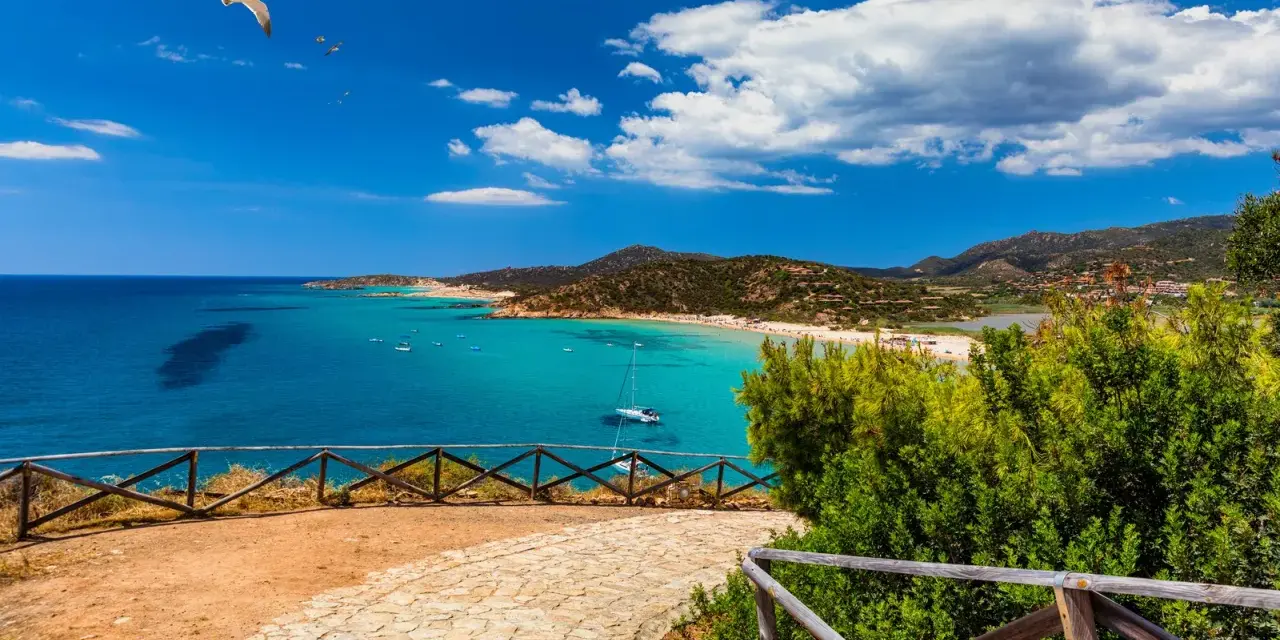 Spiaggia da sogno in Sardegna sud, cosa vedere: mare turchese, sabbia bianca, colline verdi e un gabbiano in volo.