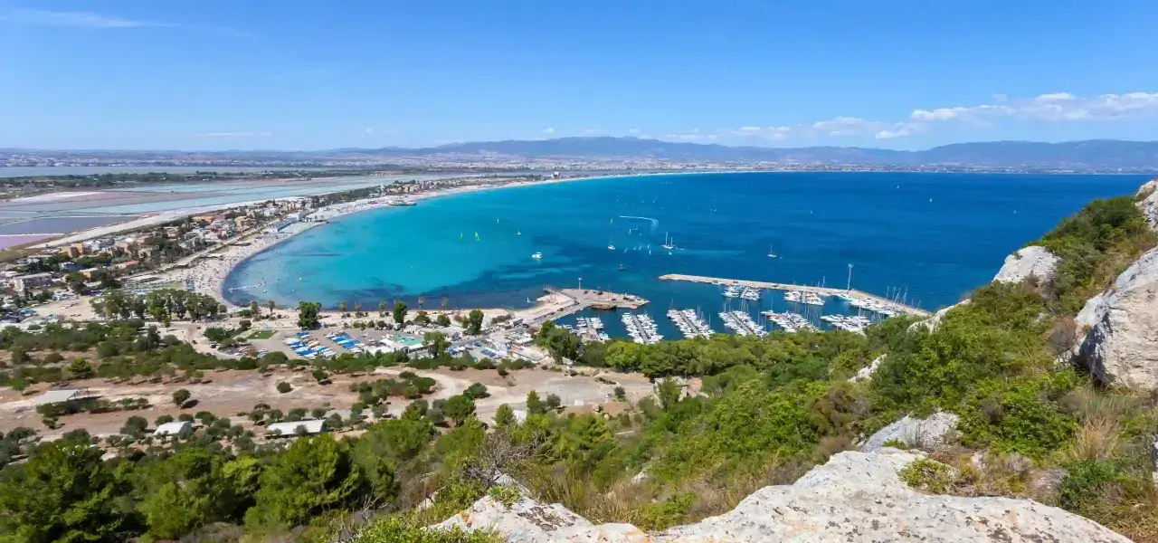 Vista panoramica della spiaggia del Poetto con mare turchese, porto turistico e saline rosa sullo sfondo.
