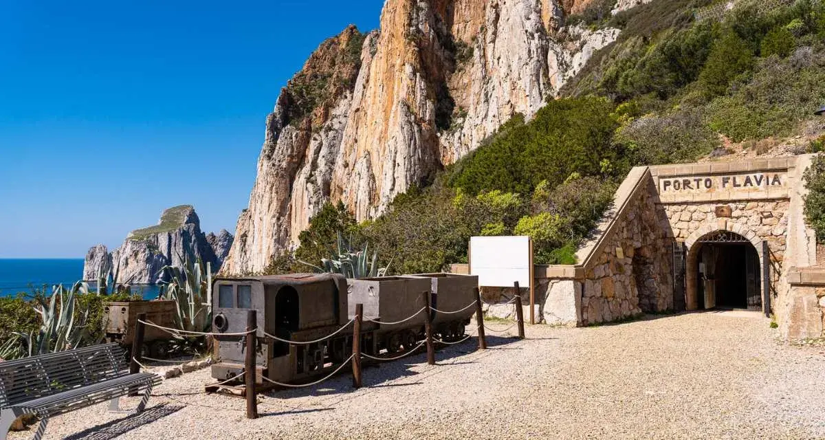 Antichi vagoni minerari e l'ingresso di Porto Flavia, testimoni della storica zona mineraria sarda, si stagliano contro un imponente promontorio roccioso.