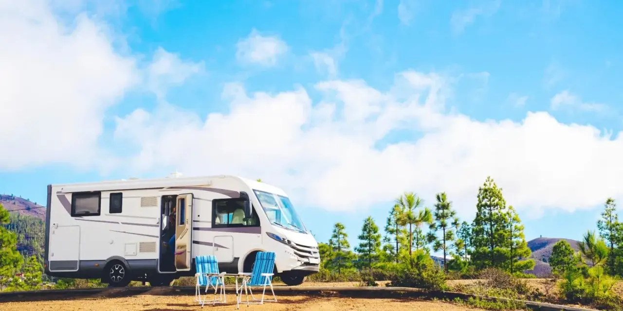 Un camper bianco è parcheggiato in un'area sterrata con due sedie blu e un tavolino. Sullo sfondo, pini e colline sotto un cielo azzurro con nuvole bianche. Un luogo ideale per un soggiorno in stile piscin'e janas.