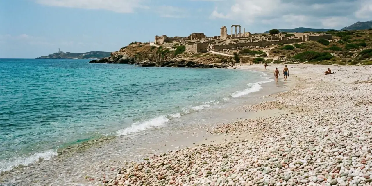 Spiaggia di ciottoli con rovine antiche in collina e un faro in lontananza. Una coppia passeggia sulla riva, godendosi la brezza marina. La facella cabras illumina il paesaggio.