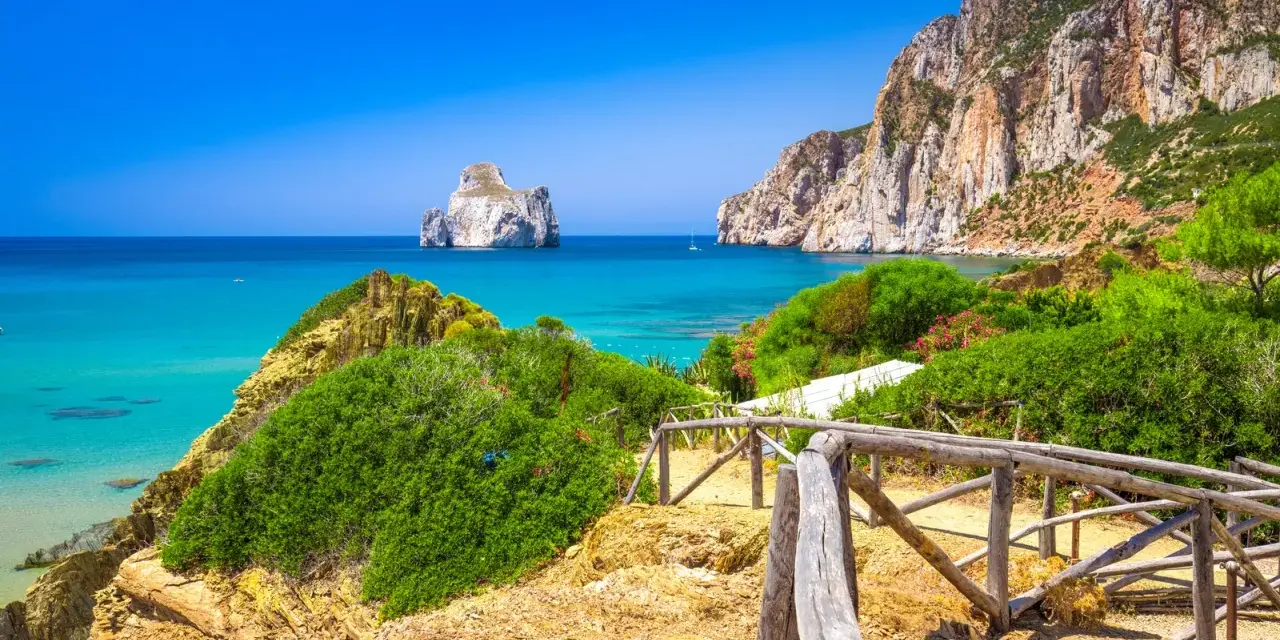 Sentiero con staccionata in legno conduce a una spiaggia con acqua cristallina e l'isolotto Pan di Zucchero in Sardegna.