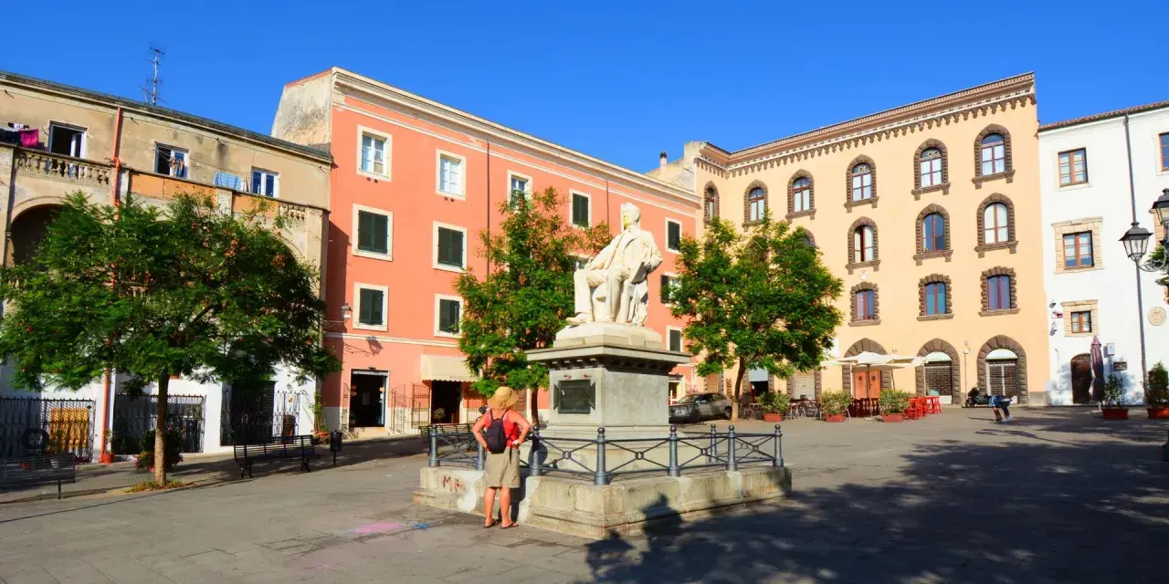 La piazza Tola a Sassari, con il monumento al centro, edifici colorati e alberi verdi sotto un cielo azzurro.