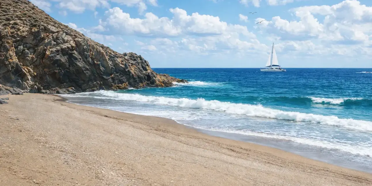 Spiaggia dorata e mare azzurro vicino a Sassari, con un catamarano bianco che naviga all'orizzonte sotto un cielo nuvoloso.