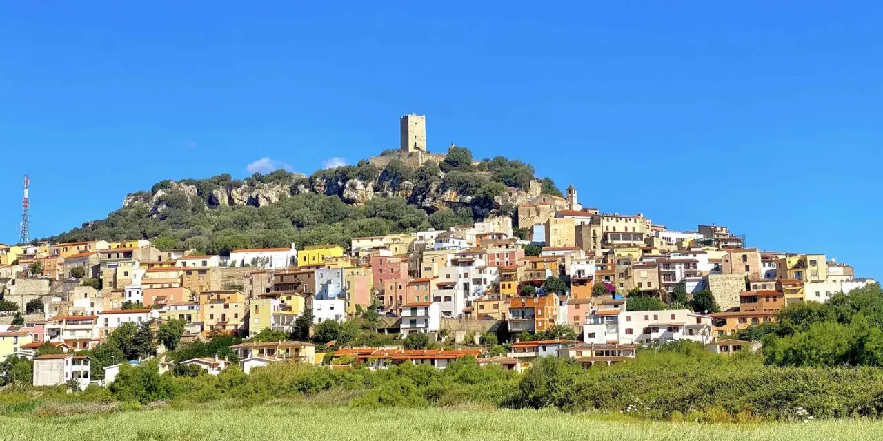 La **posada di sera** si staglia su una collina rocciosa, con case colorate e una torre medievale sotto un cielo azzurro.
