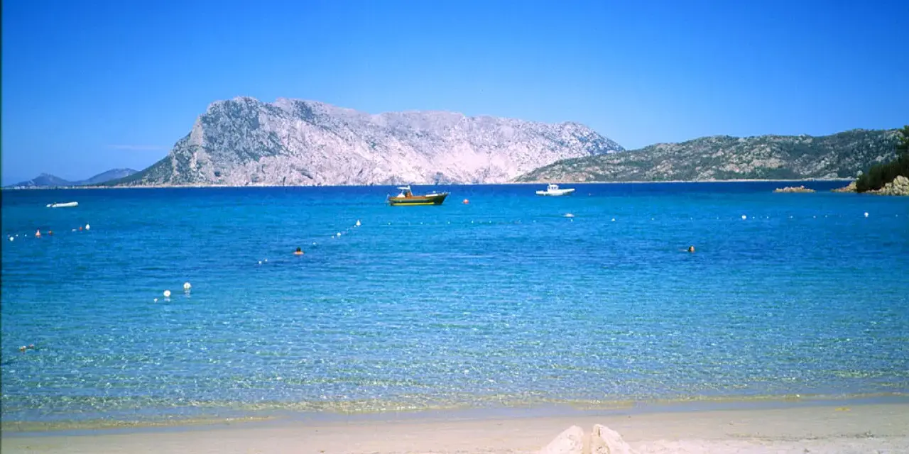 Spiaggia di Cala Suaraccia, mare cristallino, montagne sullo sfondo e barche in lontananza. Come arrivare: goditi il panorama!