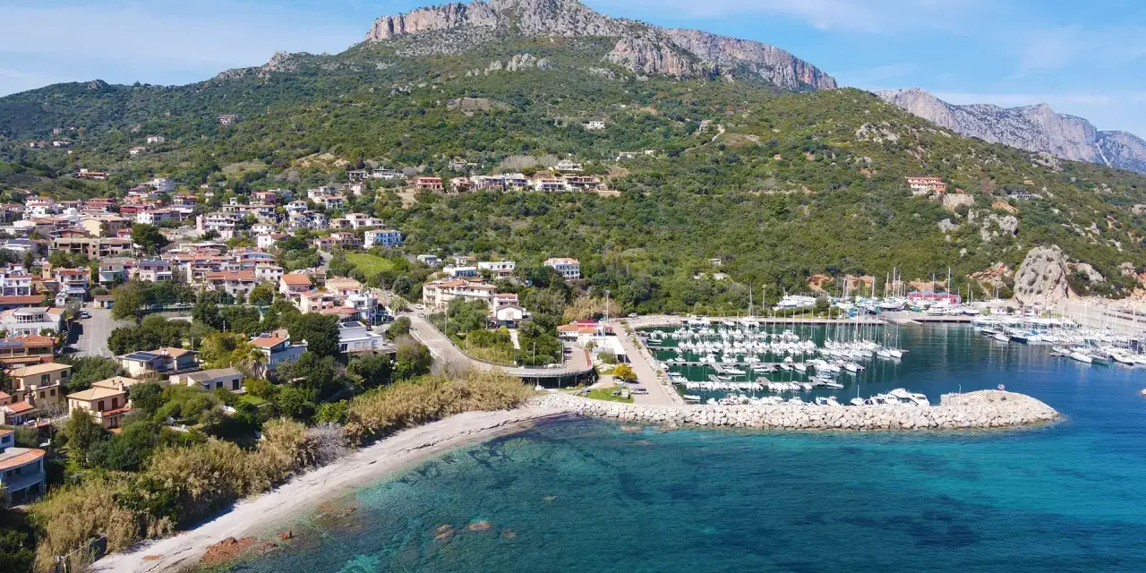 Vista aerea del centro storico di Santa Maria Navarrese, con case arroccate sulla collina, un porto turistico pieno di barche e il mare cristallino.