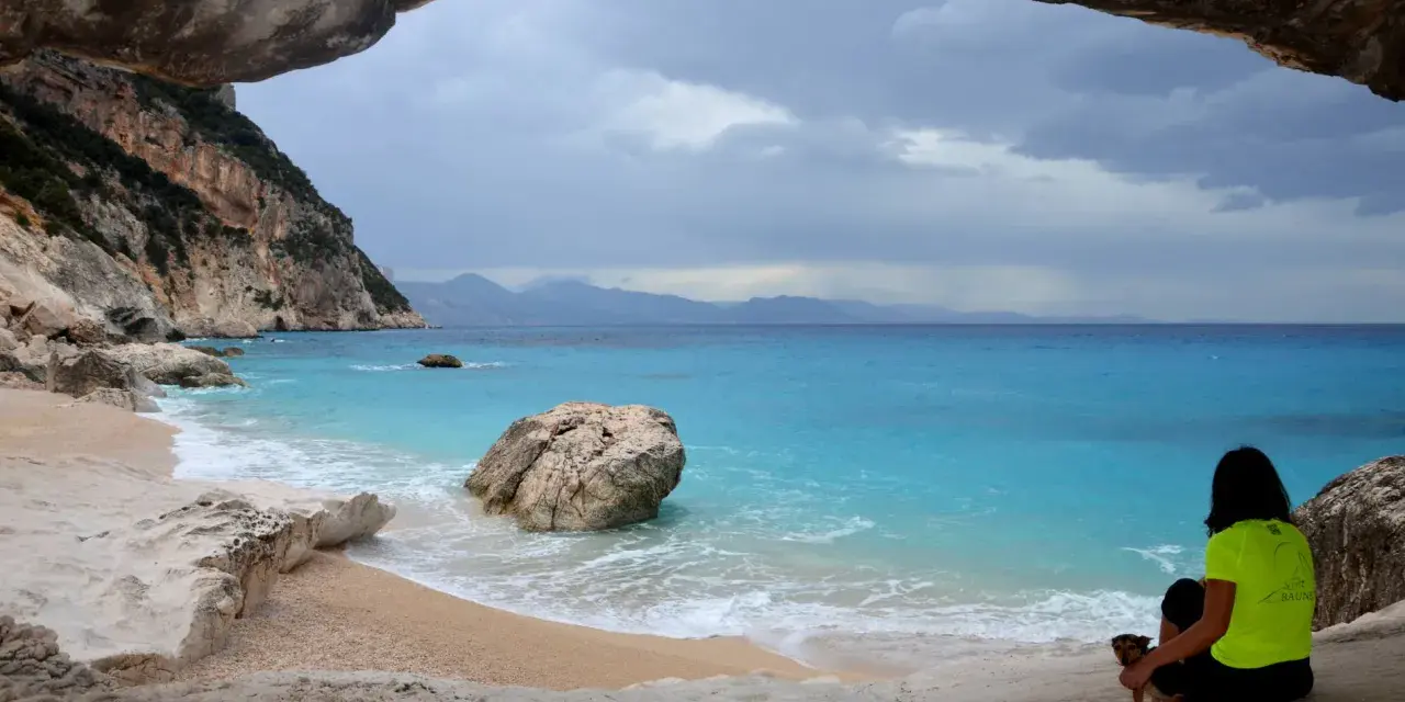 Vista da una grotta su Cala Goloritzé, con mare turchese, scogliere e una donna con un cane.