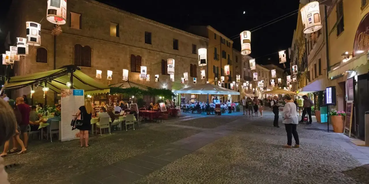 Serata ad Alghero: ristoranti all'aperto illuminati da lanterne, gente che passeggia e cena.