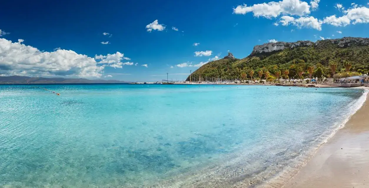 Acque cristalline e sabbia dorata sulle spiagge di Cagliari, con il promontorio verdeggiante e il porto turistico sullo sfondo.