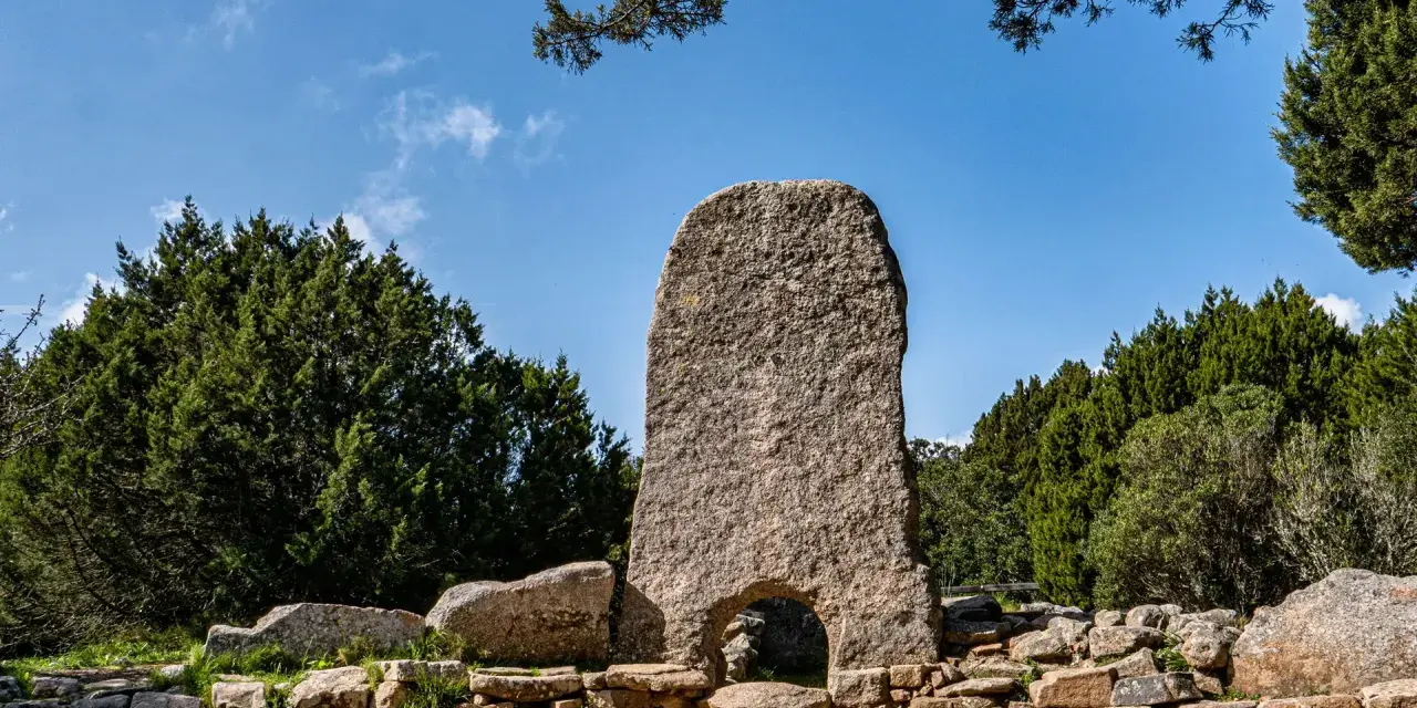 La tomba dei giganti in Sardegna, un imponente monolito di granito con un arco, circondato da pietre antiche e vegetazione rigogliosa sotto un cielo azzurro.