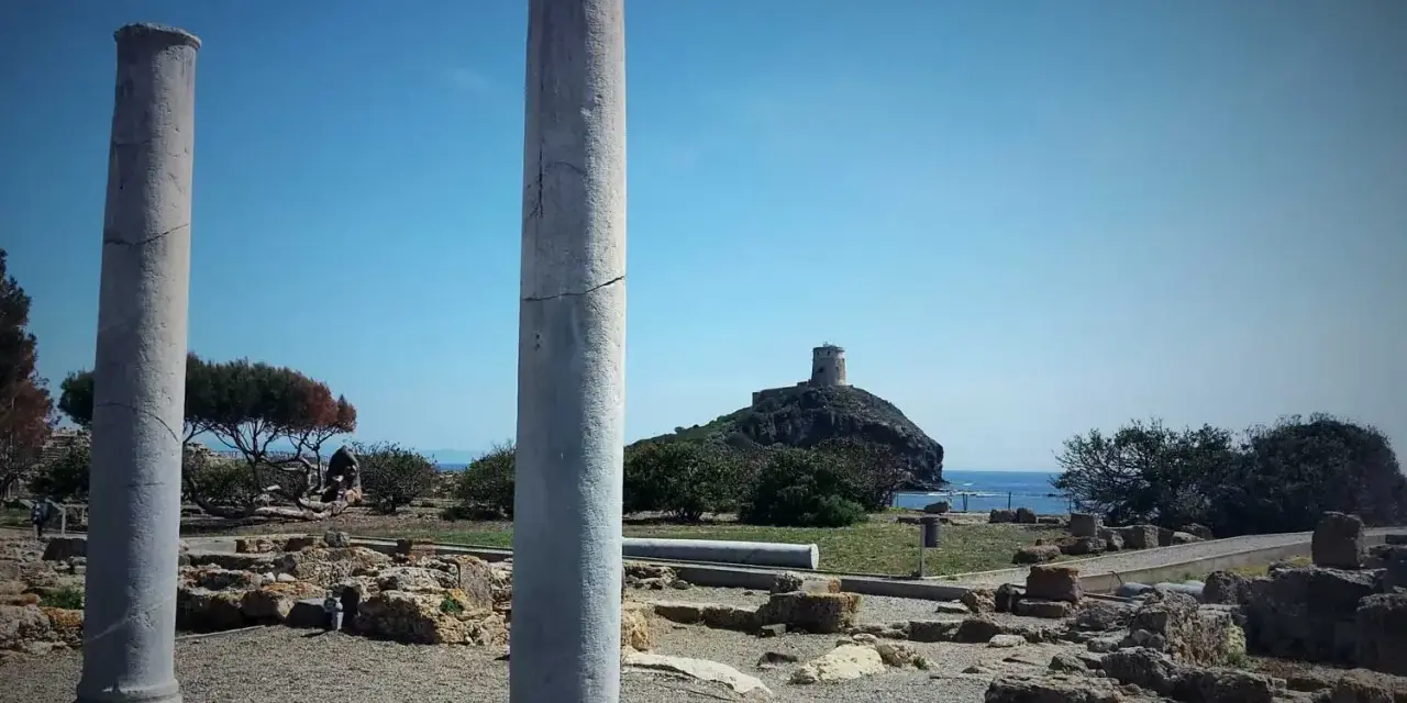 Colonne antiche nel **sito archeologico** di Nora, con una torre in lontananza su un'isola rocciosa.