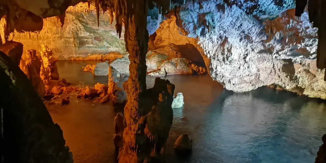 Interno di una grotta con stalattiti e stalagmiti illuminate da luci arancioni e blu. L'acqua scura riflette le formazioni rocciose in questa grotta verde.