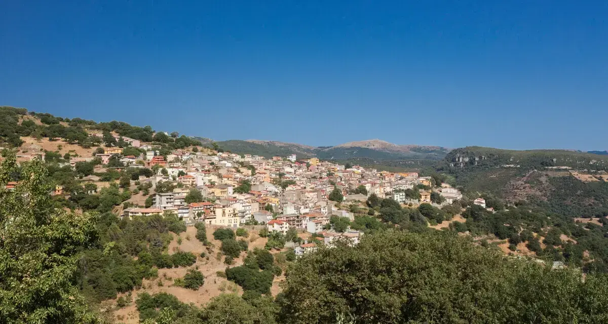 Panoramica di Seulo, Sardegna, con case colorate arroccate su una collina verdeggiante sotto un cielo azzurro.