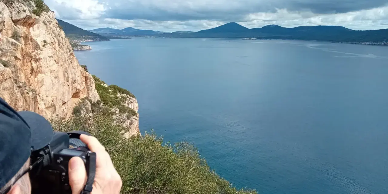 Scogliera a picco sul mare blu, con un fotografo che cattura il paesaggio. Si intravede la costa e le montagne all'orizzonte, forse vicino alla grotta verde di Alghero.