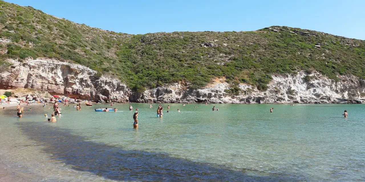 Persone si godono il sole e il mare cristallino nella spiaggia cala lunga, con colline verdi e rocce a fare da cornice.